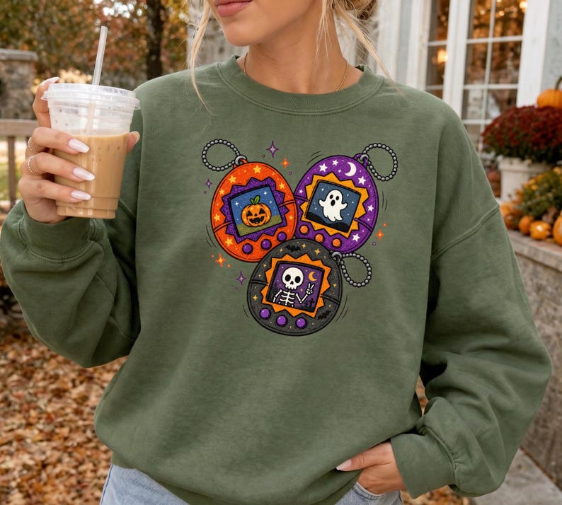 May include: An olive green crewneck sweatshirt featuring a graphic print of three digital pet keychains. The orange, purple, and black devices display a jack-o'-lantern, a white ghost, and a skeleton respectively, accented by small stars and moons.