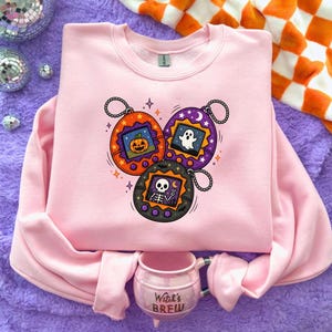 May include: A light pink crewneck sweatshirt featuring a graphic print of three digital pet keychains in orange, purple, and black. The screens display a jack-o'-lantern, a ghost, and a skeleton. Below the sweatshirt sits a pink mug with the text Witch's Brew.