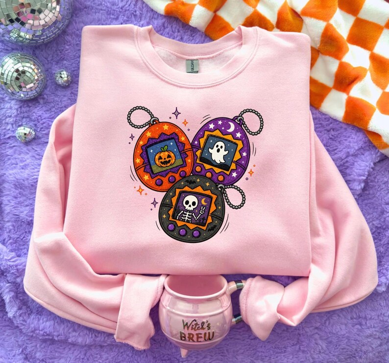 May include: A light pink crewneck sweatshirt featuring a graphic print of three digital pet keychains in orange, purple, and black. The screens display a jack-o'-lantern, a ghost, and a skeleton. Below the sweatshirt sits a pink mug with the text Witch's Brew.