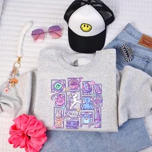 May include: A gray sweatshirt with a graphic print of a variety of 90s and early 2000s nostalgic items, including a Tamagotchi, a troll doll, a unicorn, a rainbow, and a cassette tape.