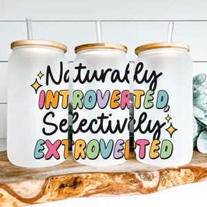 May include: Three frosted glass tumblers with bamboo lids and clear straws. The tumblers have the text "Naturally Introverted, Selectively Extroverted" in colorful bubble letters. The tumblers are on a wooden surface.
