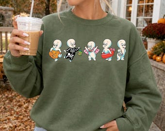 May include: An olive green crewneck sweatshirt featuring a graphic print of five cartoon ghosts in various Halloween costumes. The ghosts are depicted holding a jack-o'-lantern, wearing a skeleton suit, dressed as a vampire, using a pool float, and carrying a candy bucket.