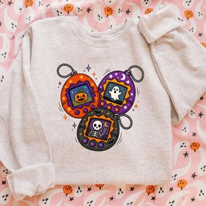 May include: A light gray crewneck sweatshirt featuring a graphic print of three oval-shaped digital pet toys. The orange toy displays a jack-o'-lantern, the purple toy shows a ghost, and the black toy depicts a skeleton making a peace sign.