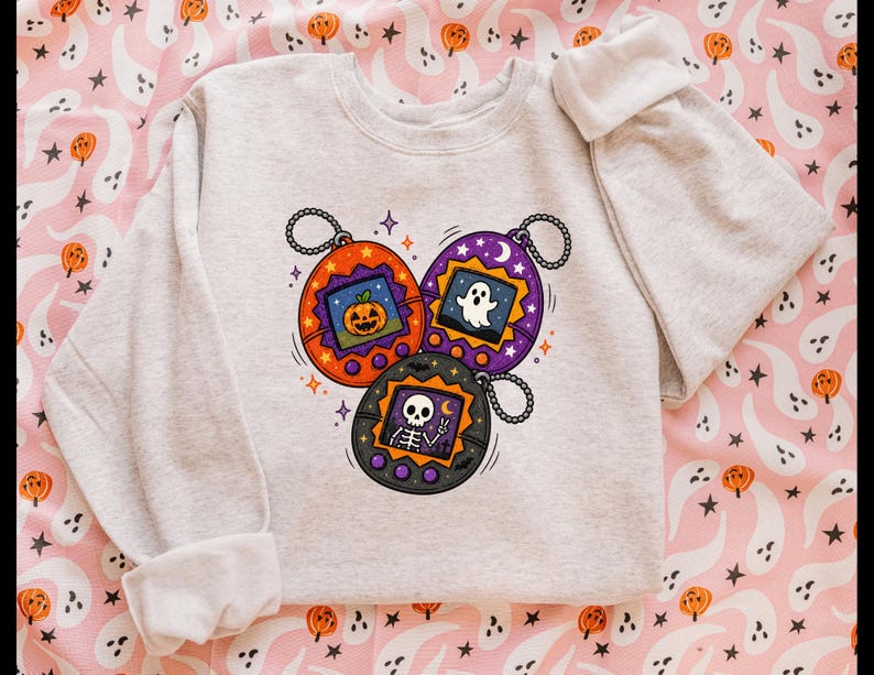 May include: A light gray crewneck sweatshirt featuring a graphic print of three oval-shaped digital pet toys. The orange toy displays a jack-o'-lantern, the purple toy shows a ghost, and the black toy depicts a skeleton making a peace sign.