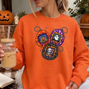 May include: An orange crewneck sweatshirt featuring a graphic print of three digital pet toys. The orange toy displays a jack-o'-lantern, the purple toy shows a white ghost, and the black toy depicts a skeleton making a peace sign gesture.