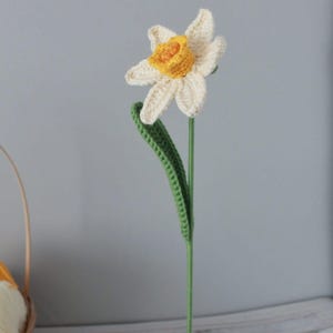 May include: A crocheted daffodil flower with a white and yellow flower and green leaves. The flower is attached to a green stem.