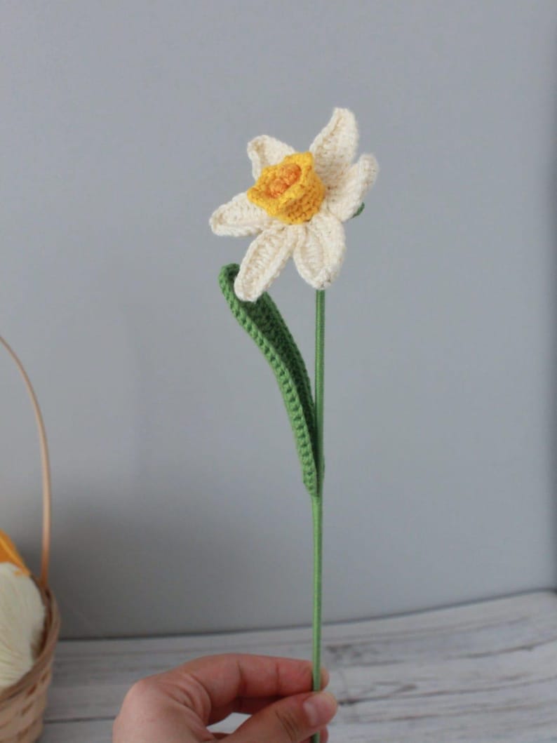 May include: A crocheted daffodil flower with a white and yellow flower and green leaves. The flower is attached to a green stem.