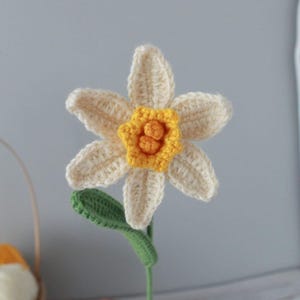 May include: A crocheted daffodil flower with a yellow center and white petals. The flower is attached to a green stem.