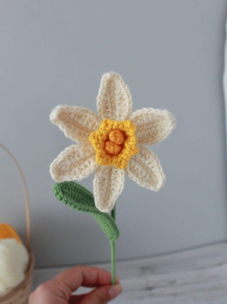 May include: A crocheted daffodil flower with a yellow center and white petals. The flower is attached to a green stem.