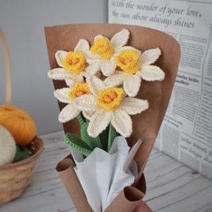 May include: A bouquet of crocheted daffodils in a brown paper wrapper with white tissue paper. The flowers are yellow and white with green stems.