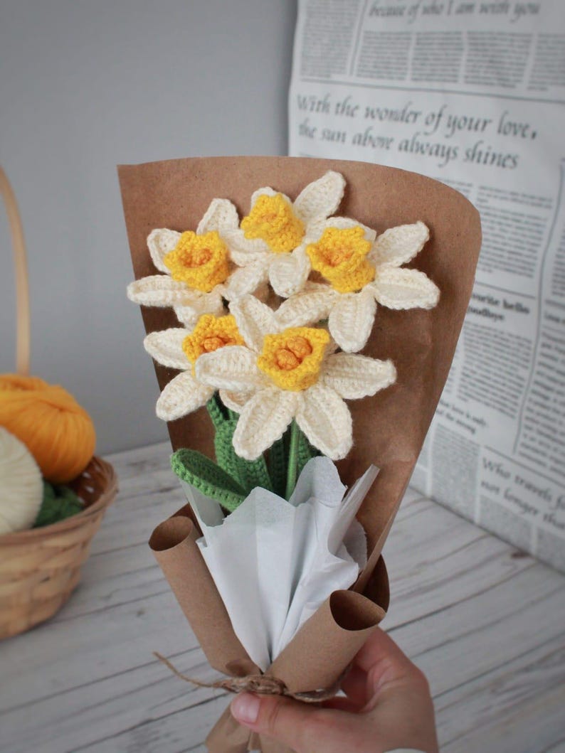 May include: A bouquet of crocheted daffodils in a brown paper wrapper with white tissue paper. The flowers are yellow and white with green stems.