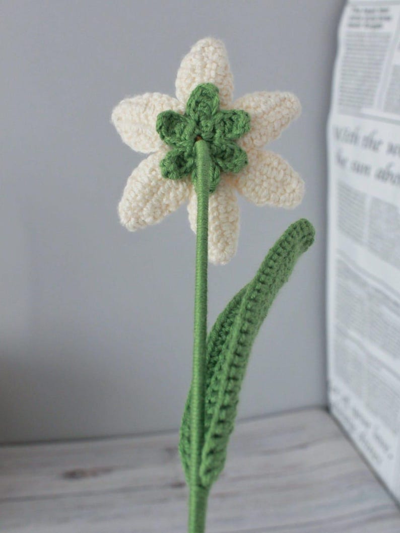 May include: A crocheted white flower with green leaves and a stem. The flower has six petals and a green center.
