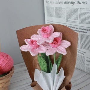 May include: A bouquet of pink crocheted flowers wrapped in brown paper with white tissue paper. The flowers are arranged in a vase and have green stems.