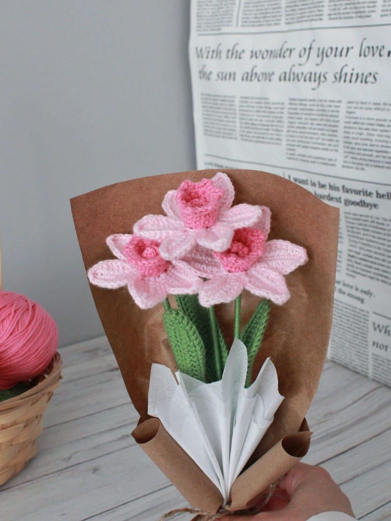 May include: A bouquet of pink crocheted flowers wrapped in brown paper with white tissue paper. The flowers are arranged in a vase and have green stems.