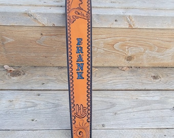 May include: A brown leather gun sling with a black border and a detailed design. The sling has a fish and moose design and the name "FRANK" is embossed in blue lettering.