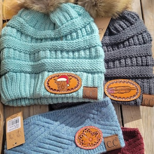 May include: Four knitted hats with faux fur pom poms. The hats are in various colors, including blue, gray, and burgundy. Each hat has a leather patch with a different design, including a cow, a feather, a flower, and a horse. The hats have the text "CC" on the leather patches.