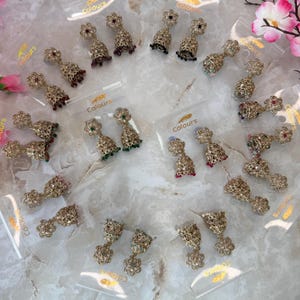 May include: A collection of gold-toned, ornate earrings with various colored gemstones, including red, green, and black. Each earring is packaged in a clear plastic bag with the word "Colours" printed on it. The earrings have a floral design.