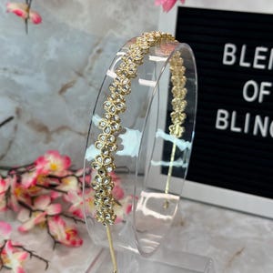 May include: A clear plastic headband with a gold floral design. The flowers are adorned with clear, sparkling accents. The headband is displayed on a clear stand, with the text "BLEND OF BLING" in the background.