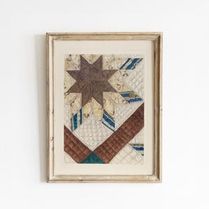 May include: Framed vintage quilt art featuring a central brown star design. The quilt has a patchwork pattern with cream, blue, and brown fabrics. The artwork is encased in a distressed, light-colored wooden frame, adding a rustic touch to the piece.