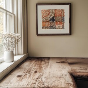 May include: A framed artwork with a patchwork design in shades of orange, white, and navy blue hangs on a beige wall. A rustic wooden countertop with a natural finish is in the foreground, near a white window frame and a vase of dried flowers.