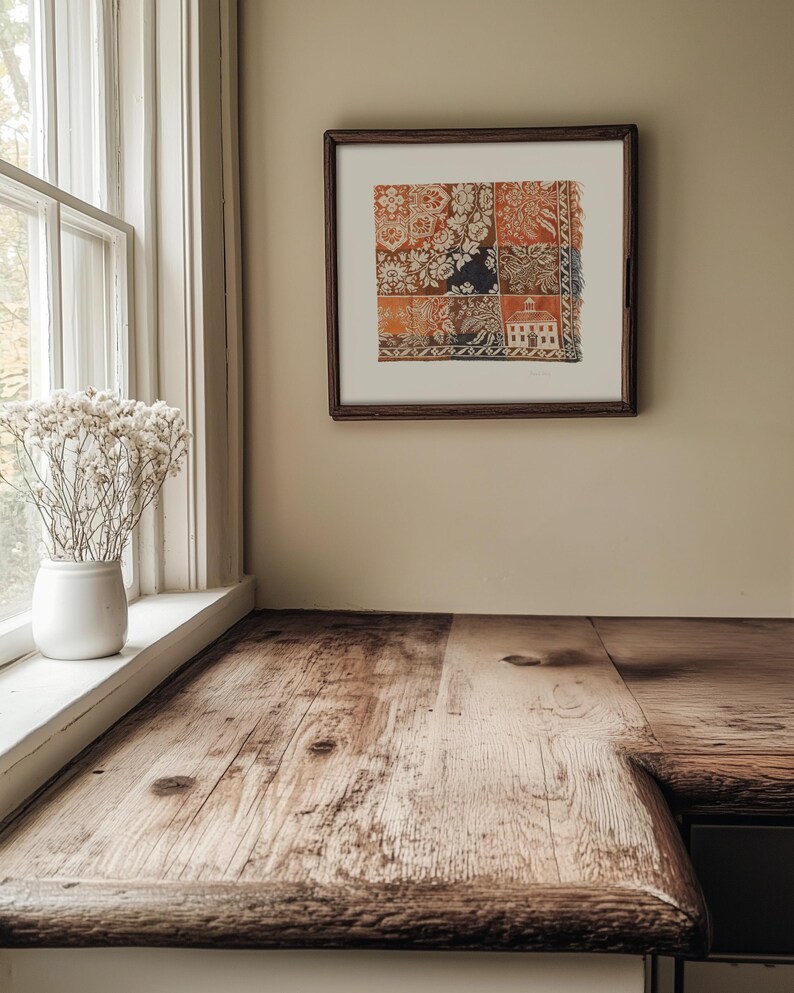 May include: A framed artwork with a patchwork design in shades of orange, white, and navy blue hangs on a beige wall. A rustic wooden countertop with a natural finish is in the foreground, near a white window frame and a vase of dried flowers.