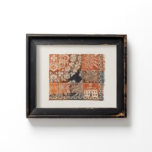 May include: Framed artwork featuring a patchwork design with floral and architectural motifs in shades of orange, white, brown, and navy blue. The artwork is encased in a distressed black wooden frame, creating a vintage aesthetic.