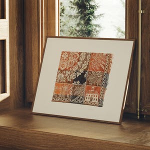 May include: A framed print featuring a detailed textile pattern with floral and geometric designs in shades of red, orange, and brown. The artwork is displayed on a wooden surface near a window, showcasing a classic aesthetic.