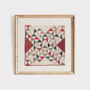 Könnte beinhalten: Gerahmte Quiltkunst mit einem Patchwork-Design aus roten, blauen und neutralen Dreiecken. Der Quilt ist in einem goldfarbenen Holzrahmen gerahmt, was eine Vintage-Ästhetik erzeugt. Das Design des Quilts ist symmetrisch und geometrisch.