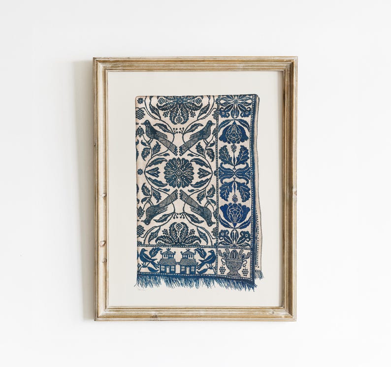 May include: Framed print of a blue and white textile featuring a detailed floral and bird design. The pattern includes stylized birds, flowers, and foliage, with a fringed edge. The artwork is presented in a light-coloured wooden frame.