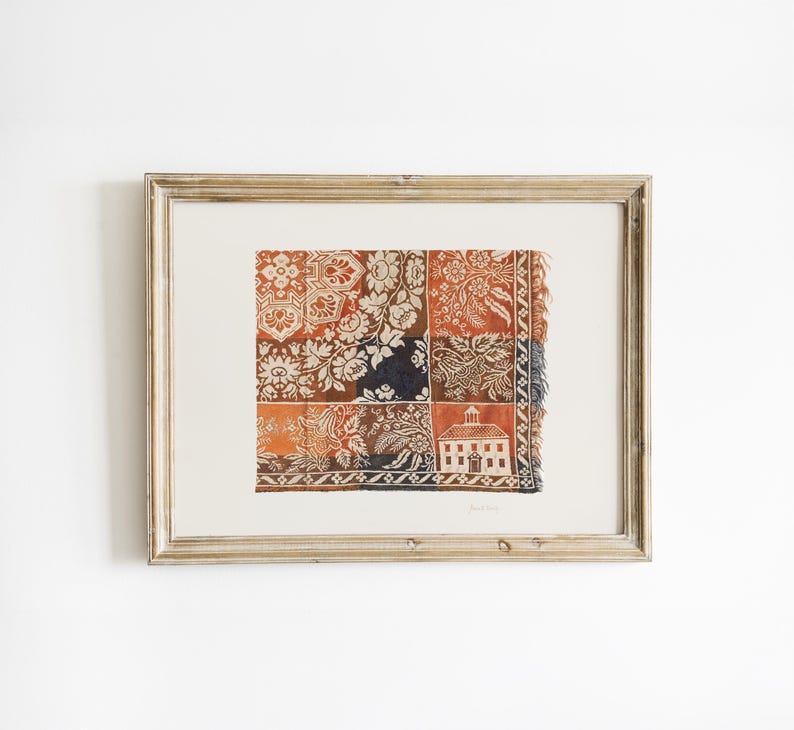 May include: Framed artwork with a patchwork design in orange, brown, and white, featuring floral and geometric patterns. A small white building illustration is included. The frame is light brown.