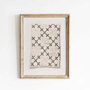 Puede incluir: Obra de arte textil enmarcada con una tela color crema con un patrón de cruces negras repetidas. La tela tiene una apariencia texturizada y está enmarcada en un marco de madera dorada. La obra textil tiene el número 1824 bordado.