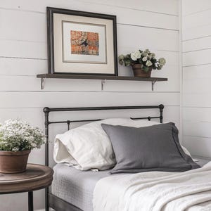 May include: A bedroom scene featuring a bed with white and gray bedding, including pillows and a blanket. A framed artwork hangs above the bed on a shelf, and a potted plant sits on a side table, creating a cozy atmosphere.