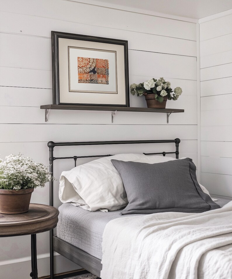 May include: A bedroom scene featuring a bed with white and gray bedding, including pillows and a blanket. A framed artwork hangs above the bed on a shelf, and a potted plant sits on a side table, creating a cozy atmosphere.