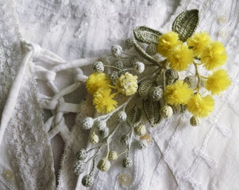 May include: A crocheted flower brooch with yellow and green accents. The brooch is pinned to a white fabric with lace detailing.