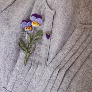 May include: A crocheted flower brooch featuring purple and yellow pansies with green leaves. The brooch is pinned to a grey fabric background.