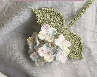 May include: A crocheted mini hydrangea with green leaves. The hydrangea is made with white, pink, and blue yarn. The text "CROCHET MINI HYDRANGEA" and "www.floralcrochet.com" is on a pink background.