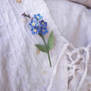 May include: A delicate crocheted forget-me-not flower brooch in shades of blue and green, pinned to a white lace fabric.