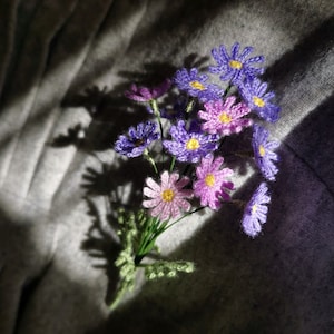 以下が含まれることがあります： 紫とピンクの色合いの、かぎ針編みで作った花束。花は黄色い中心を持っており、集まって配置されています。