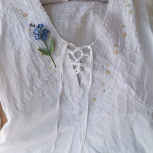 May include: White cotton blouse with a lace trim and a delicate floral embroidery design. The blouse has a V-neckline and a lace-up detail at the front. A small blue forget-me-not flower is pinned to the neckline.