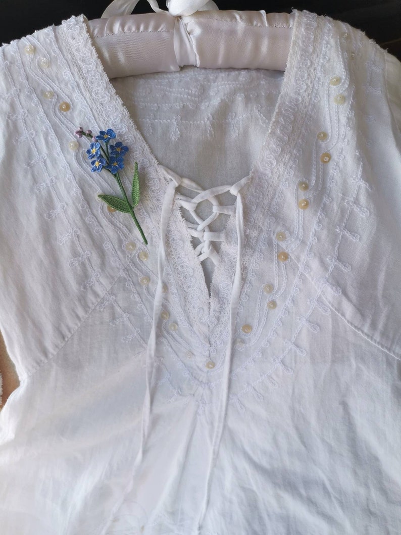 May include: White cotton blouse with a lace trim and a delicate floral embroidery design. The blouse has a V-neckline and a lace-up detail at the front. A small blue forget-me-not flower is pinned to the neckline.