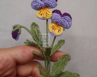 May include: A crocheted purple and yellow pansy flower with green leaves. The flower is handmade and has a delicate, realistic appearance.