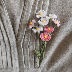 Könnte beinhalten: Ein Strauß gehäkelter Blumen in Rosa-, Weiß- und Gelbtönen mit grünen Stielen, an einem grauen Stoffhintergrund befestigt.