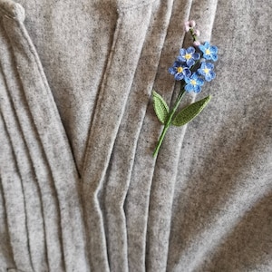 May include: A delicate crocheted forget-me-not flower brooch in shades of blue and green, pinned to a gray fabric background.