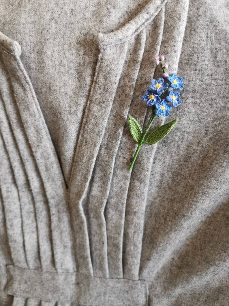 May include: A delicate crocheted forget-me-not flower brooch in shades of blue and green, pinned to a gray fabric background.