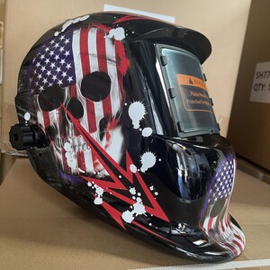 May include: A black welding helmet with a skull and American flag design. The helmet has a clear viewing window and a warning label that reads "Please Remove Protective Film Before Use".