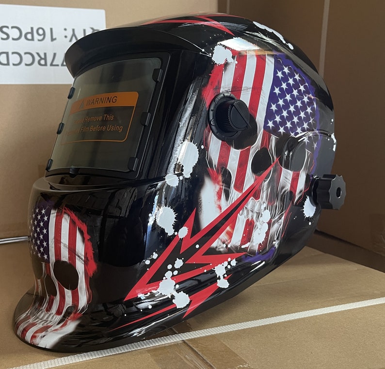 May include: A black welding helmet with a skull and American flag design. The helmet has a clear viewing window and a warning label that says "Remove This Film Before Using".