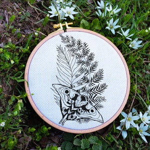 May include: A black and white embroidery hoop with a fern and a moth design. The hoop is on a bed of green grass and white flowers.