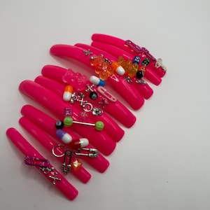 May include: Pink long false nails with various small charms, including gummy bears, pills, safety pins, and other small trinkets.