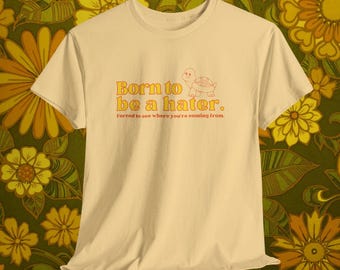 Könnte beinhalten: Cremefarbenes T-Shirt mit dem orangefarbenen Schriftzug "Born to be a hater." und einer Schildkröten-Illustration. Darunter steht der Satz "Forced to see where you're coming from." Das T-Shirt hat einen Rundhalsausschnitt und kurze Ärmel.