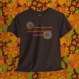 以下が含まれることがあります： 「I'm not a saint. I just prefer to get paid for my mental breakdowns」というテキストが入った濃い茶色のTシャツ。テキストはオレンジ色で、デイジーには笑顔の顔があります。Tシャツは花柄の背景にあります。
